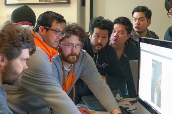 a group of programmers huddled around a computer screen, intensely discussing and brainstorming essential features for a zillow-like app development project.